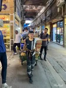 کودک خندان باربر در حال حمل چرخ بار در بازار آهنگرهای تهران