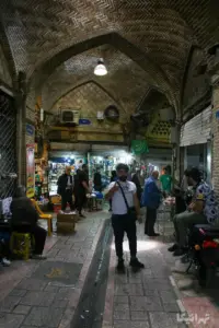 مردی در میانه بازار کوچه مروی به دوربین نگاه می‌کند و با دست علامت تایید می‌دهد