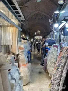 مردی جلوی طاقه‌های پارچه ایستاده و مرد دیگری درون مغازه خود در دالان سوم ملک بازار نشسته