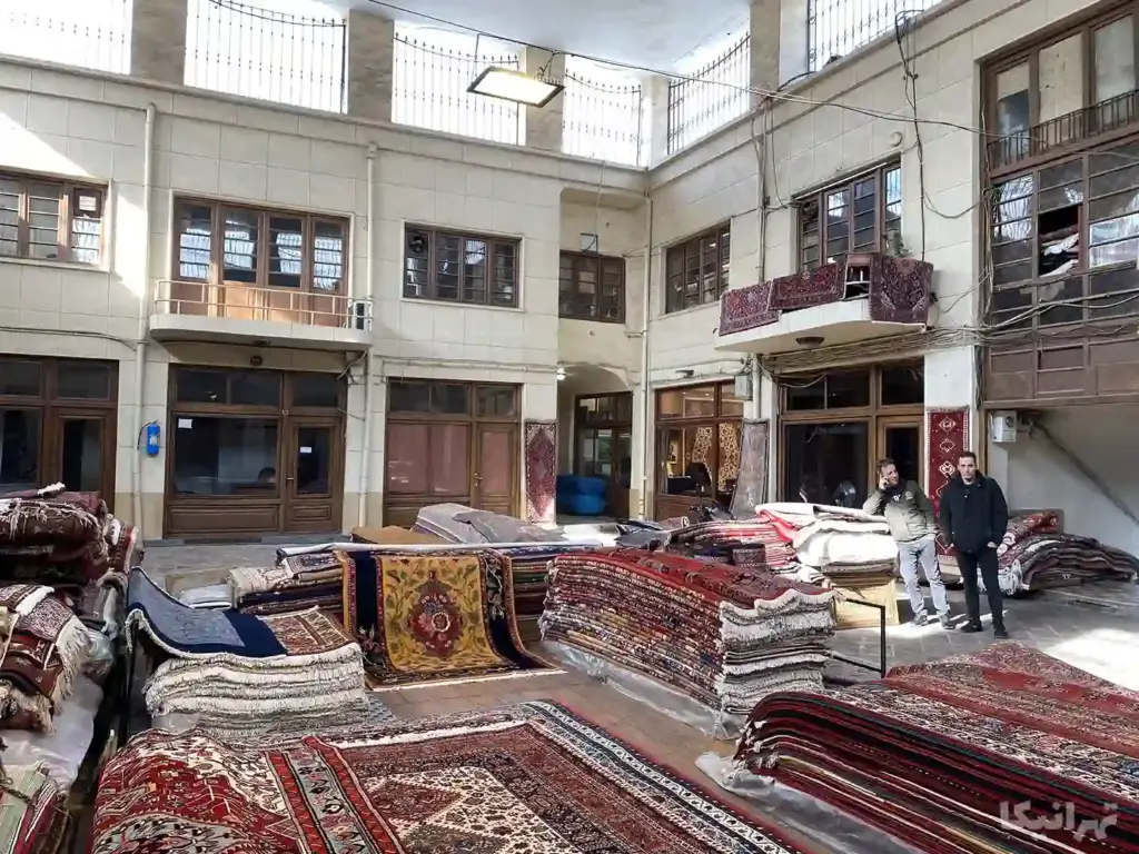 دو مرد در کنار فرش‌های دستباف چیده‌شده در وسط حیاط مسقف سرای رحیمیه اول ایستاده‌اند