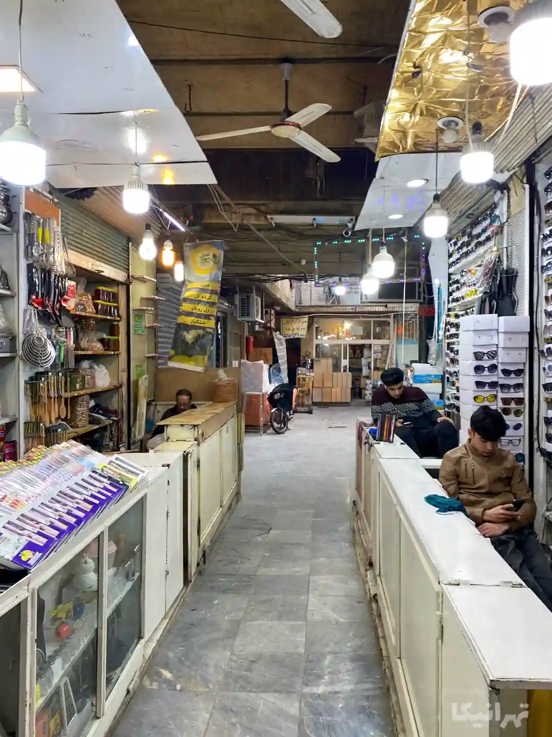 چند فروشنده پشت پیشخوان غرفه‌های دالان ورودی سرای سادات درحال کار با موبایل