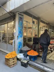 مرد دستفروش نشسته روی فرش‌ها تا شده در پشت بساط میوه خود و مردی در حال ورود به سرای قدس حسینی بازار