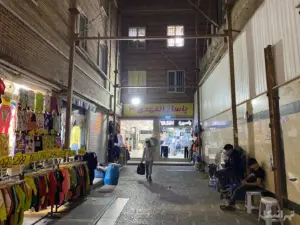 دو نفر روی صندلی کنار دیوار نشسته و مردی در حال حمل بار جلوی پاساژ المهدی ۳ بازار