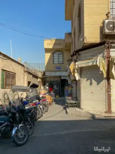 مردی در انتهای کوچه باریک در بازار تهران نشسته و مردی دیگری در حال خرید است